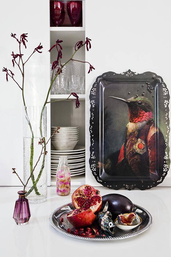 Decorative tray with bird motif, spindly twigs in vases and pomegranate in front of kitchen shelving