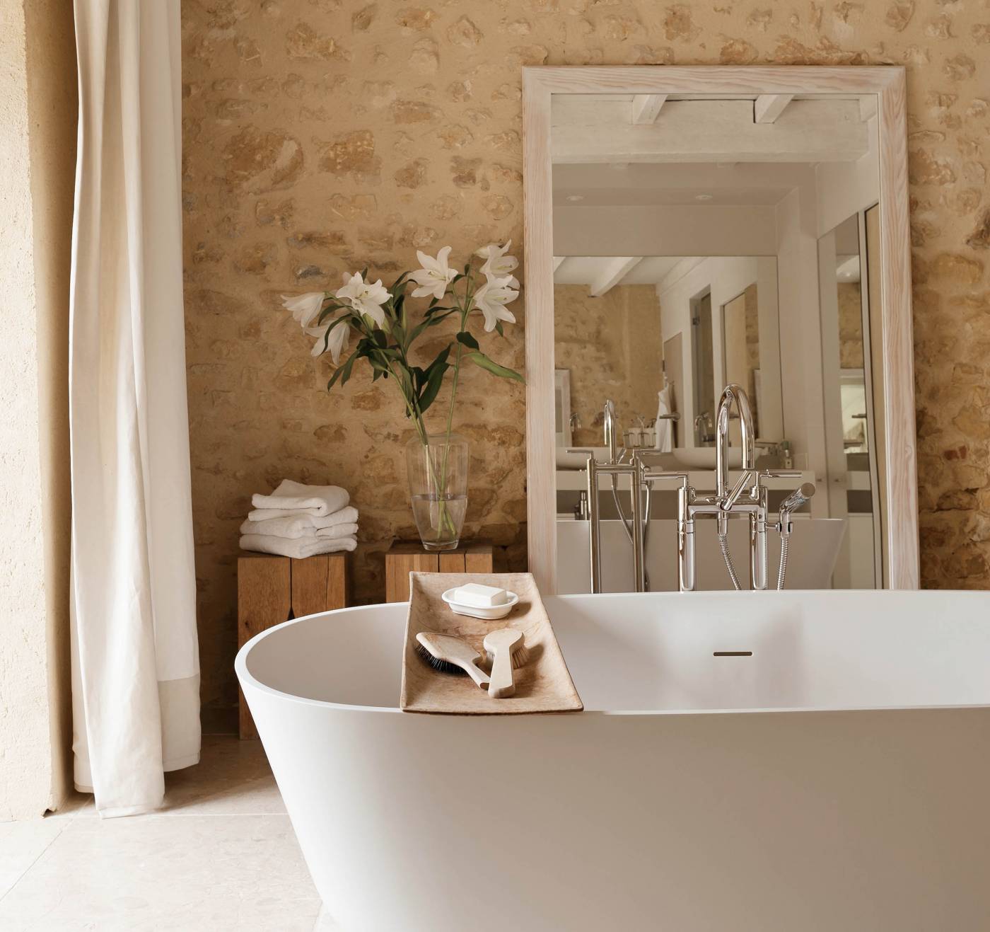 Free-standing bathtub with wooden shelf and mirror leaning against stone wall in ensuite bathroom